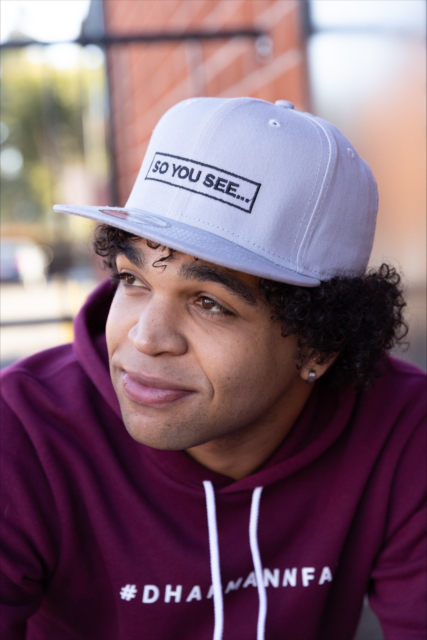 Azeem wearing the maroon DharMannFam Hoodie and the So You See... New Era snapback hat in light grey