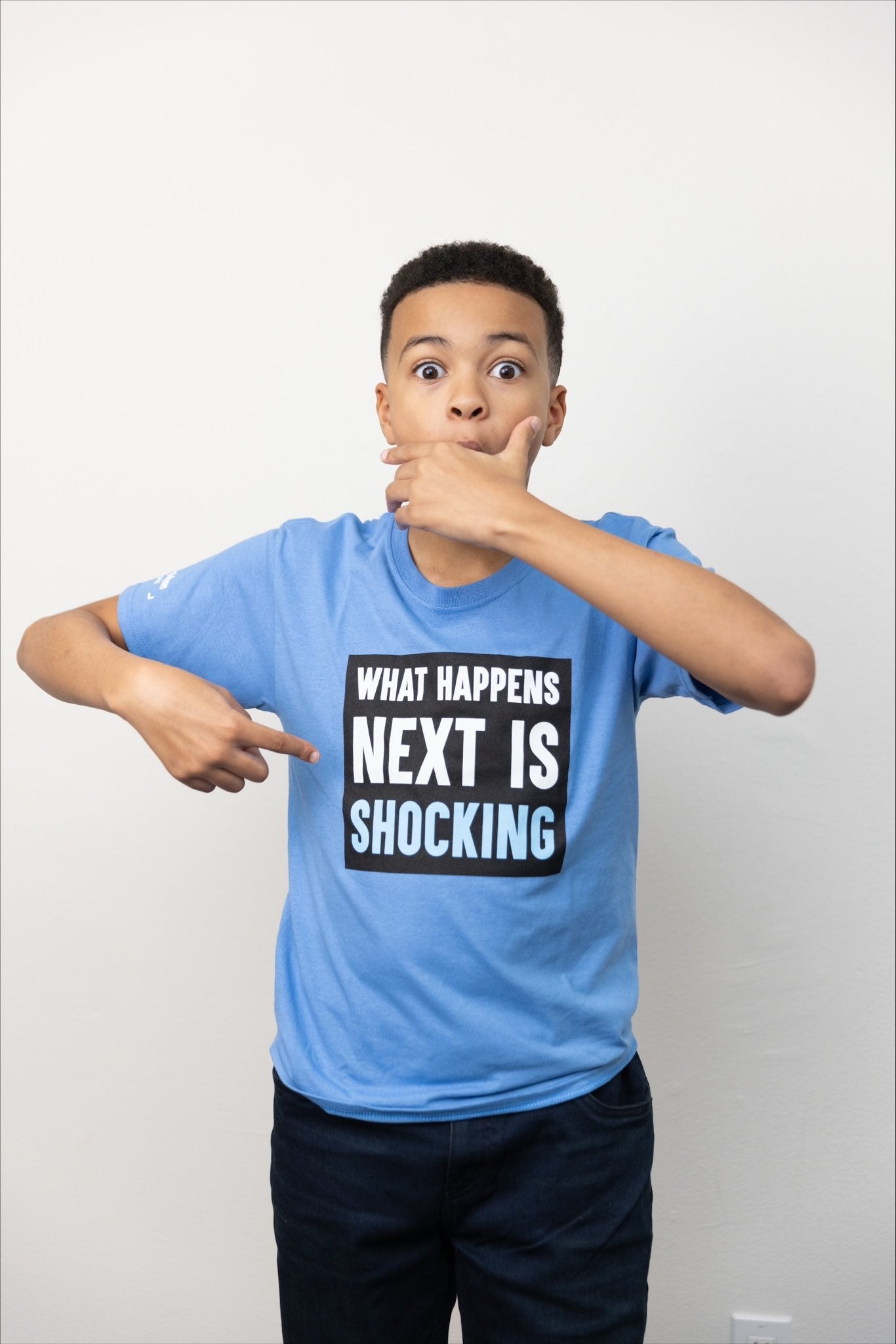 Shaun can't believe it! He's covering his mouth in disbelief and is pointing to the text on the front of his light blue t-shirt, which says, printed inside a black square:  "WHAT HAPPENS NEXT IS SHOCKING". It's truly shocking!