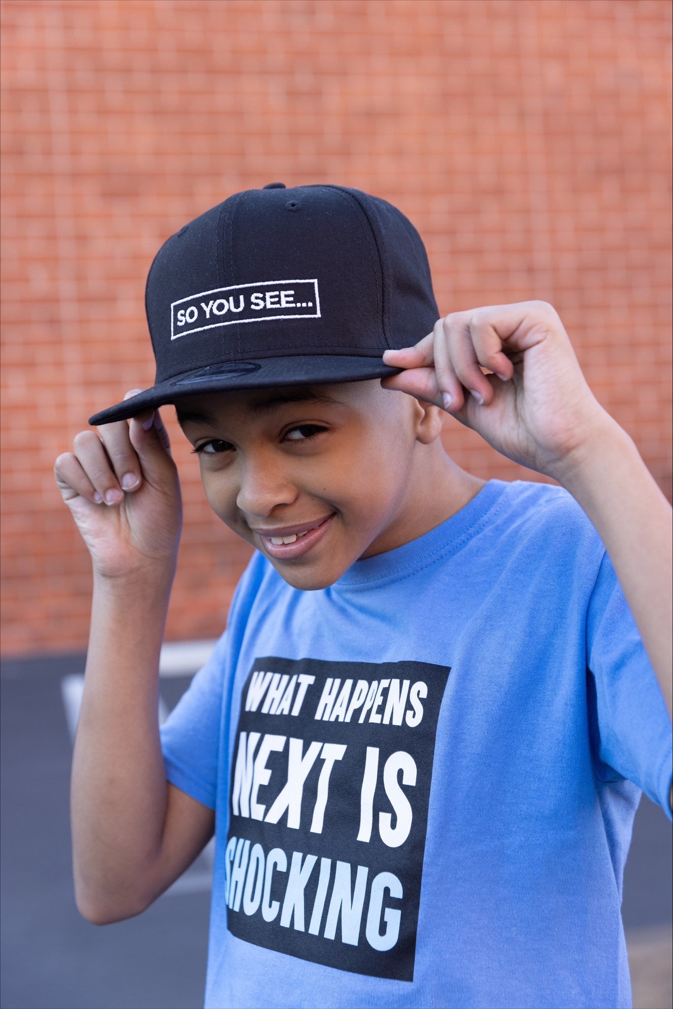 Shaun showing off his merch, adjusting the brim of his black So You See... New Era snapback cap. He is also wearing a light blue 'What Happens Next is Shocking' t-shirt. He is standing in a parking lot in front of a brick wall, likely the location of Bookside School.