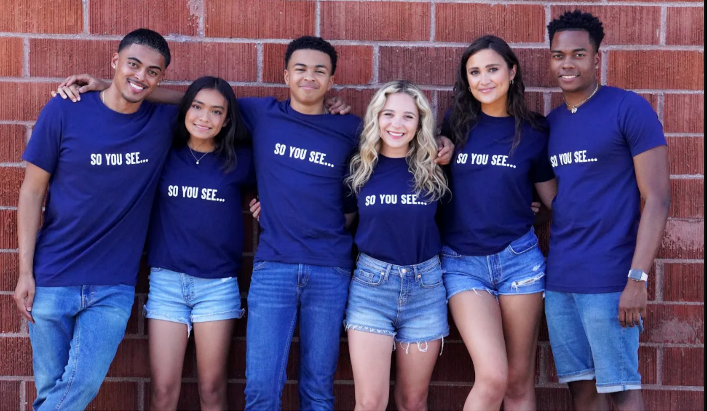 six cast-member friends lined up in front of a school's brick wall while wearing matching So You See classic Navy blue t-shirts 