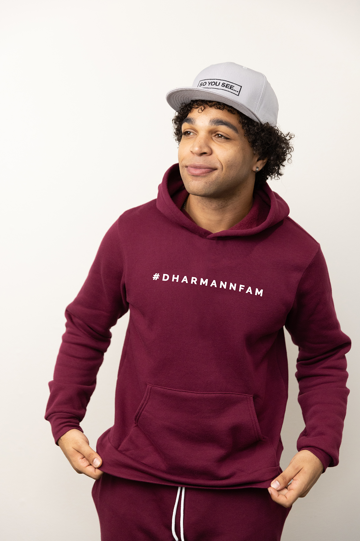 Azeem shows off his Dhar Mann Studios merch, wearing the maroon DharMannFam Hoodie and matching DharMannFam Jogger sweatpants, also in Maroon. He is also wearing the So You See... New Era snapback hat in light grey