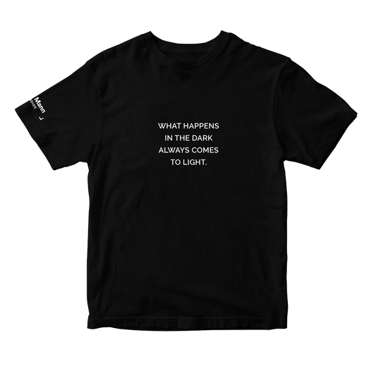 Black short sleeve t-shirt with the text "WHAT HAPPENS IN THE DARK ALWAYS COMES TO LIGHT." printed in white. It also features a printed tag on the right sleeve: the 'Dhar Mann Studios' logo. 